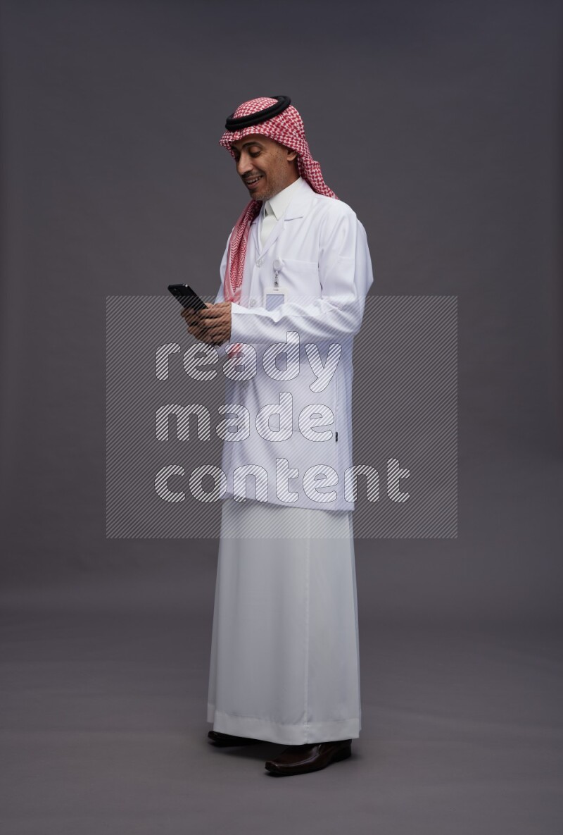Saudi man wearing thob with lab coat and shomag with pocket employee badge standing texting on phone on gray background