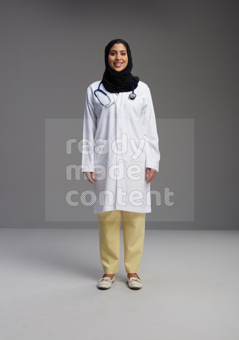 Saudi woman wearing lab coat with stethoscope standing interacting with the camera on Gray background