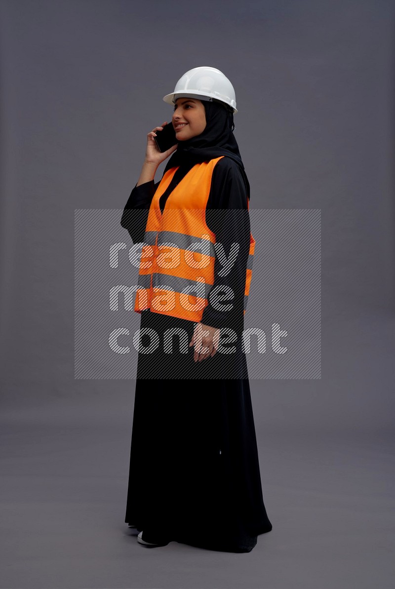 Saudi woman wearing Abaya with engineer vest standing talking on phone on gray background