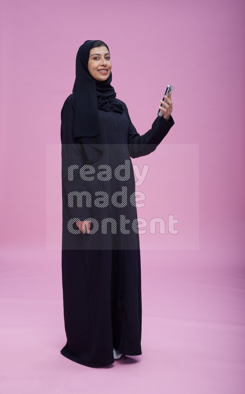 Saudi woman wearing Abaya standing taking selfie on pink background