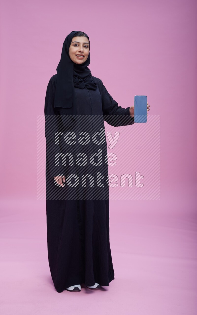 Saudi woman wearing Abaya standing showing phone to camera on pink background