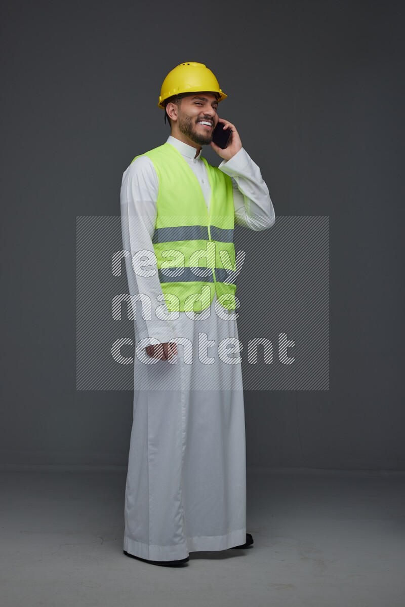 A Saudi man wearing Thobe with a yellow safety vest and white helmet standing and talking in the phone eye level on a gray background