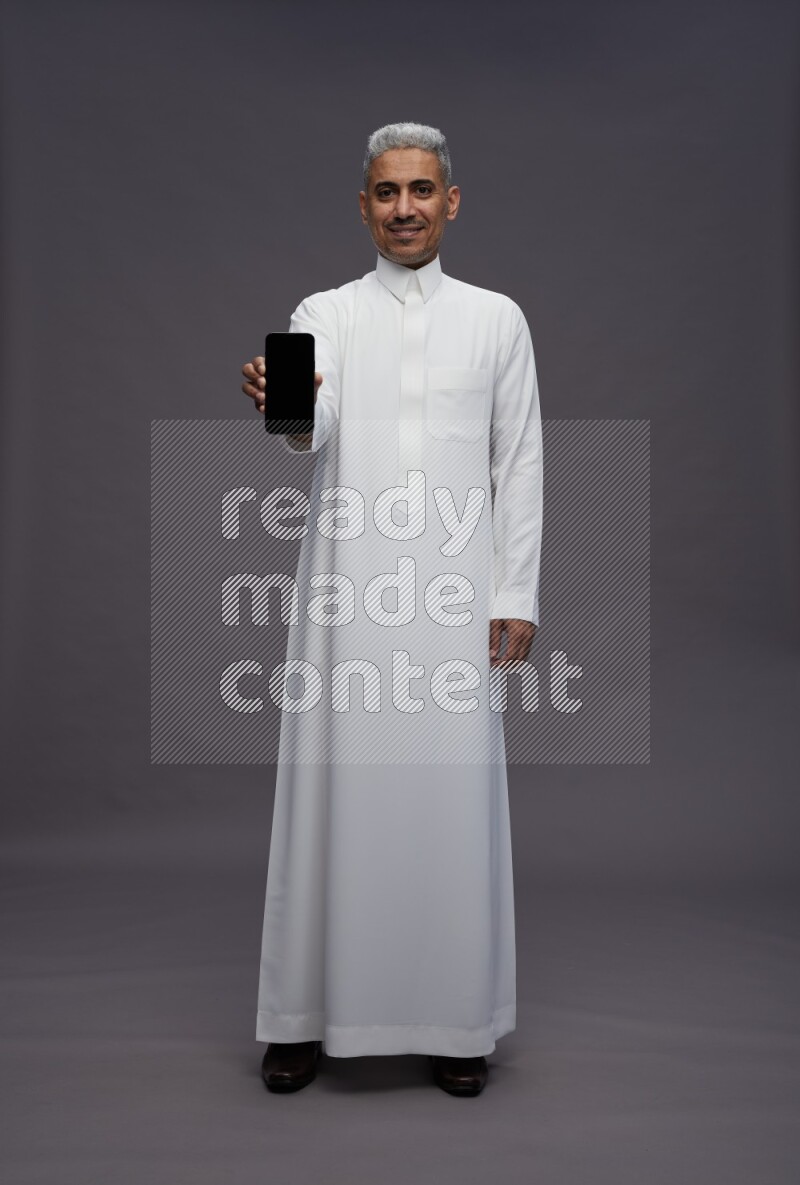 Saudi man wearing thob standing showing phone to camera on gray background