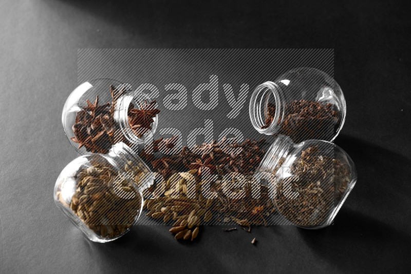 4 glass spice jars full of cloves, cardamom, basil and star anise on black flooring