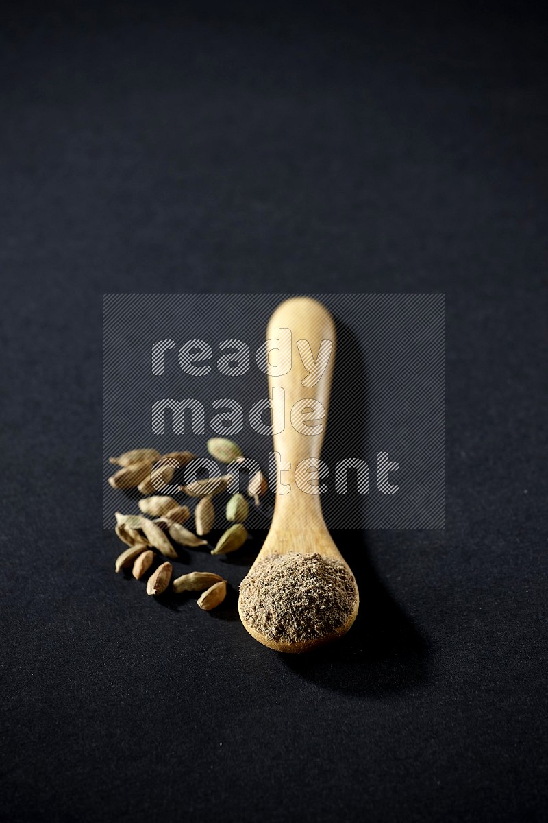 A wooden spoon full of cardamom powder and cardamom seeds beside it on black flooring