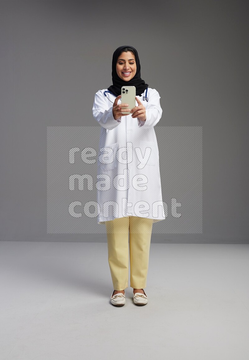 Saudi woman wearing lab coat with stethoscope standing taking selfie on Gray background