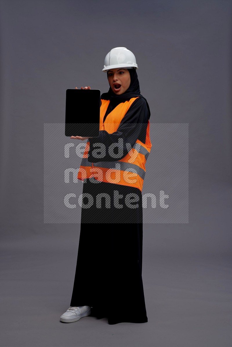 Saudi woman wearing Abaya with engineer vest standing showing tablet to camera on gray background