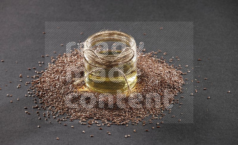 A glass jar full of flaxseeds oil surrounded by the seeds on a black flooring