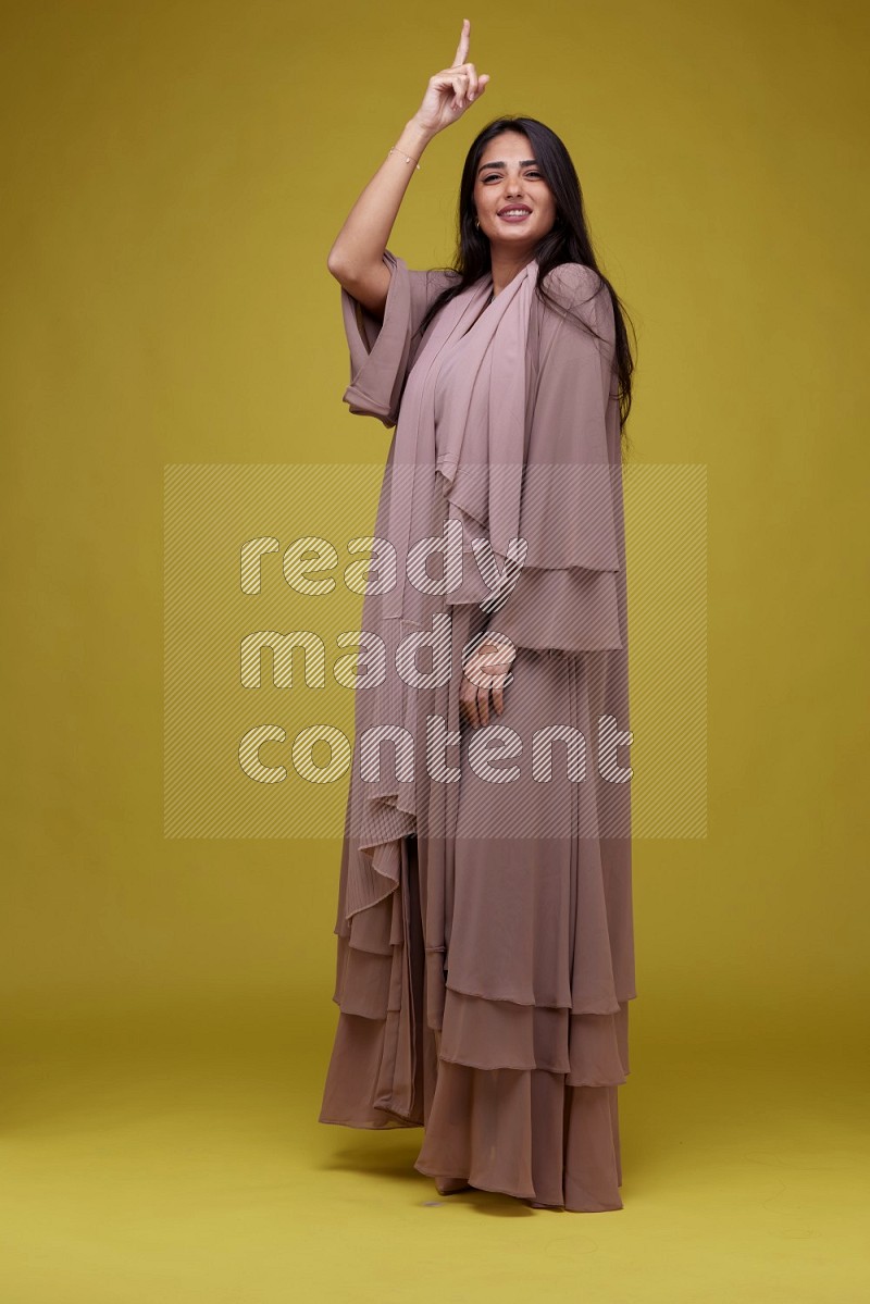 A woman Pointing on a Yellow Background wearing Brown Abaya