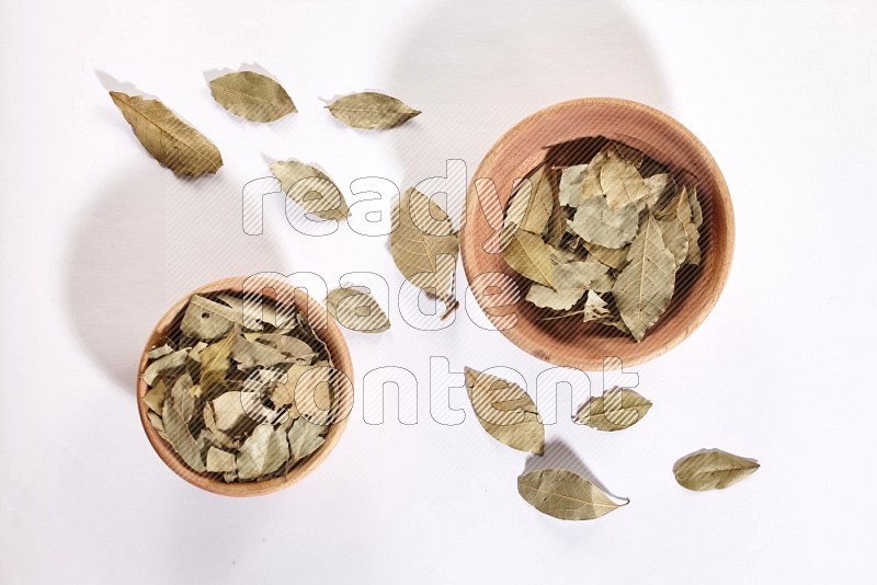 2 wooden bowls full of dried bay leaves with more leaves spread on white flooring