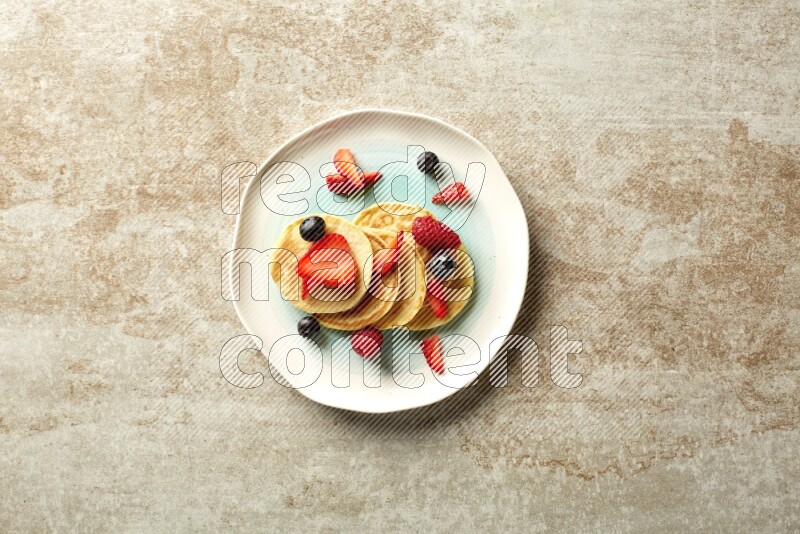 Five stacked mixed berries mini pancakes in a bicolor plate on beige background