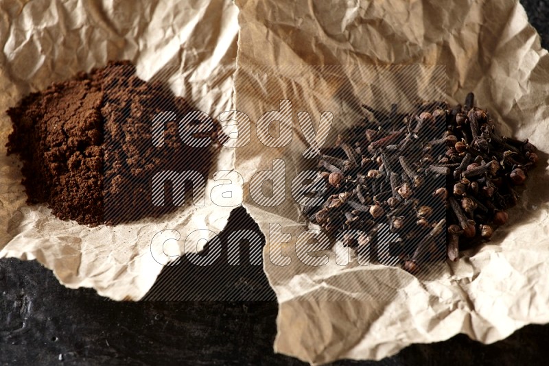 Cloves powder and cloves in 2 crumpled pieces of paper on a textured black flooring