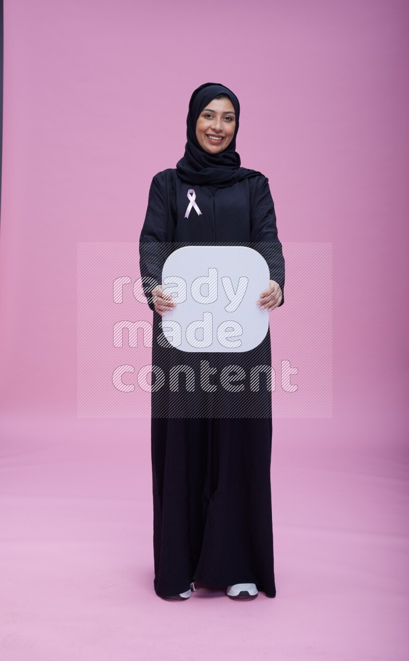 Saudi woman wearing pink ribbon on Abaya standing holding social media signs on pink background