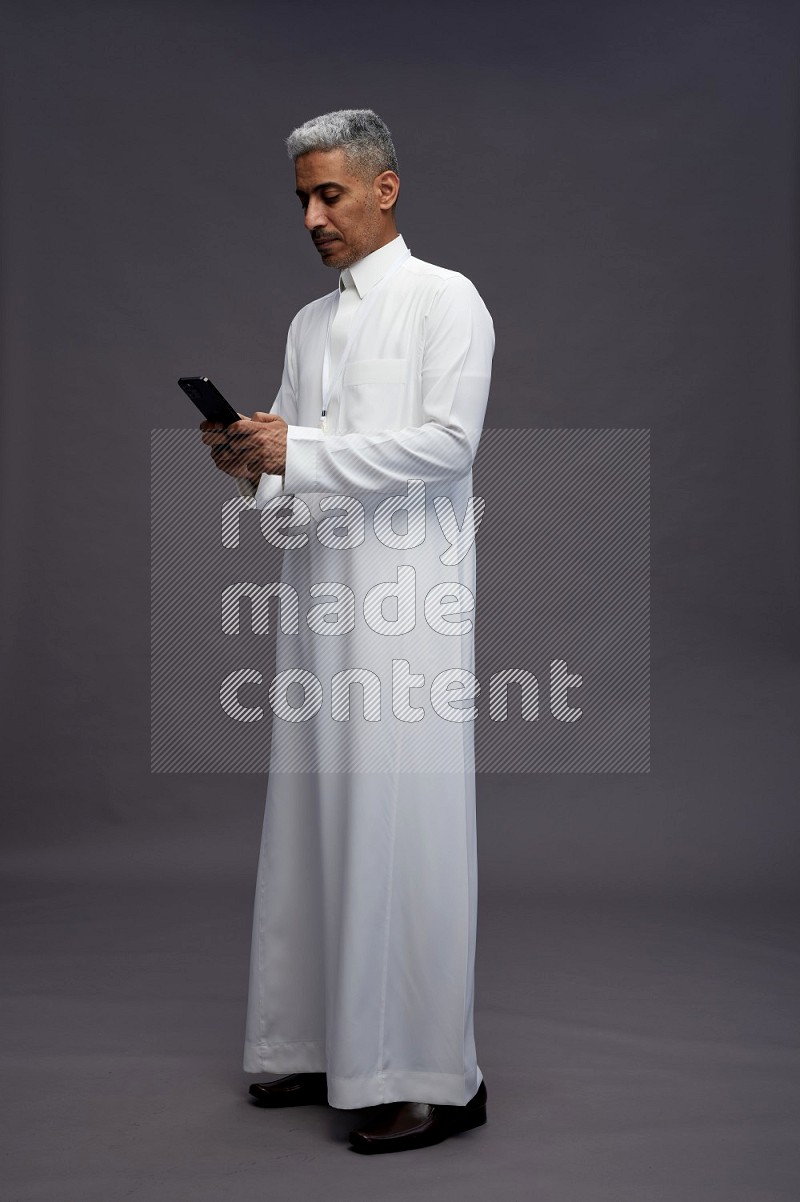 Saudi man wearing thob with neck strap employee badge standing texting on phone on gray background