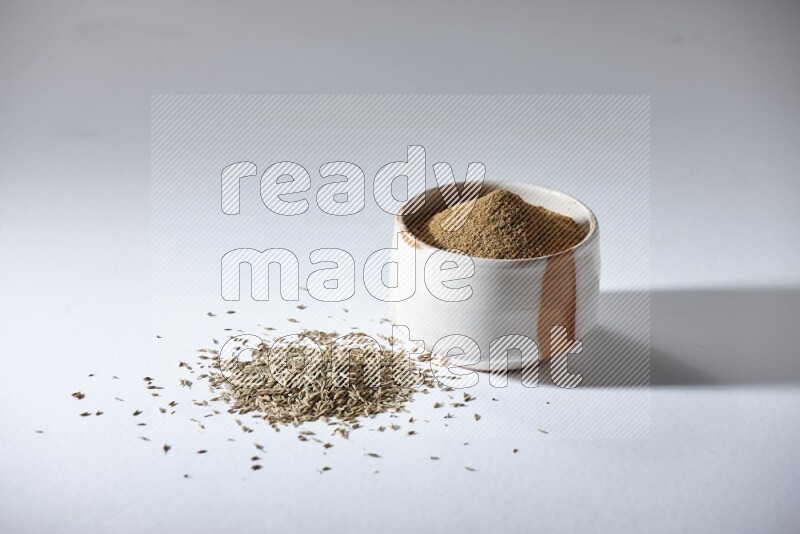 A beige bowl full of cumin powder with cumin seeds beneath it the bowl on a white flooring