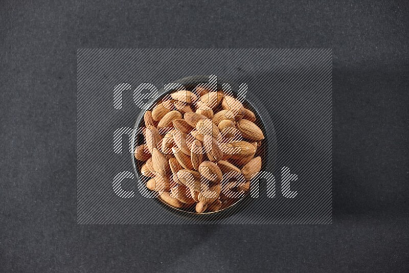 A black pottery bowl full of peeled almonds on a black background in different angles