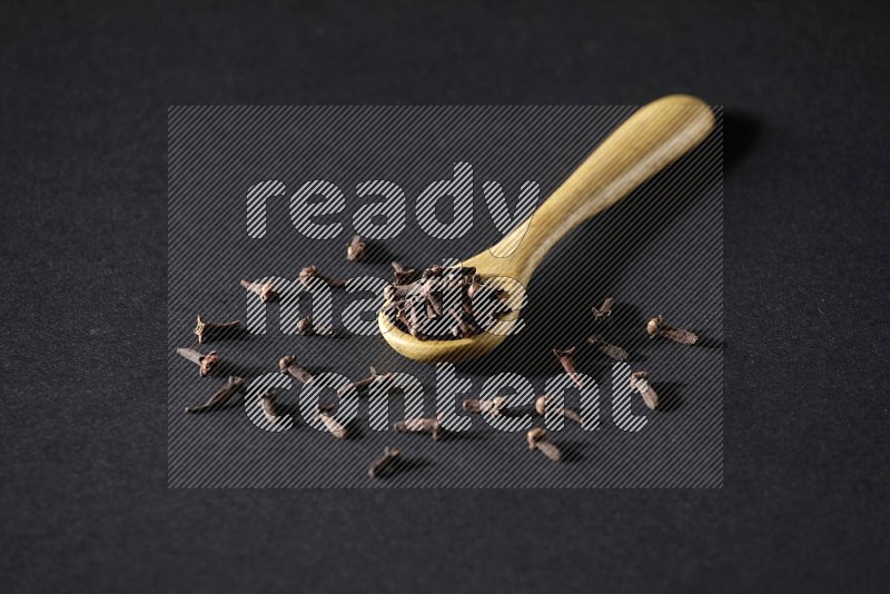 A wooden spoon full of cloves and some spreaded out of it on a black flooring
