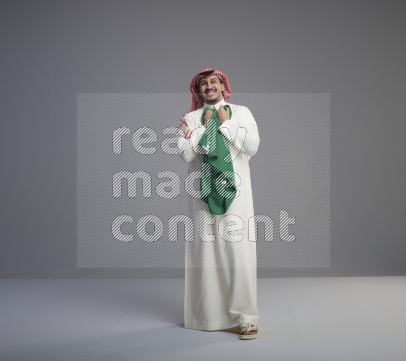 A saudi man standing wearing thob and red shomag holding big saudi flag on gray background