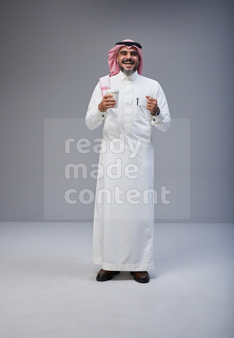 Saudi man Wearing Thob and red Shomag standing holding paper cup on Gray background