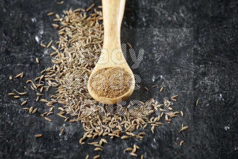 A wooden spoon full of cumin powder and cumin seeds spreaded on textured black flooring