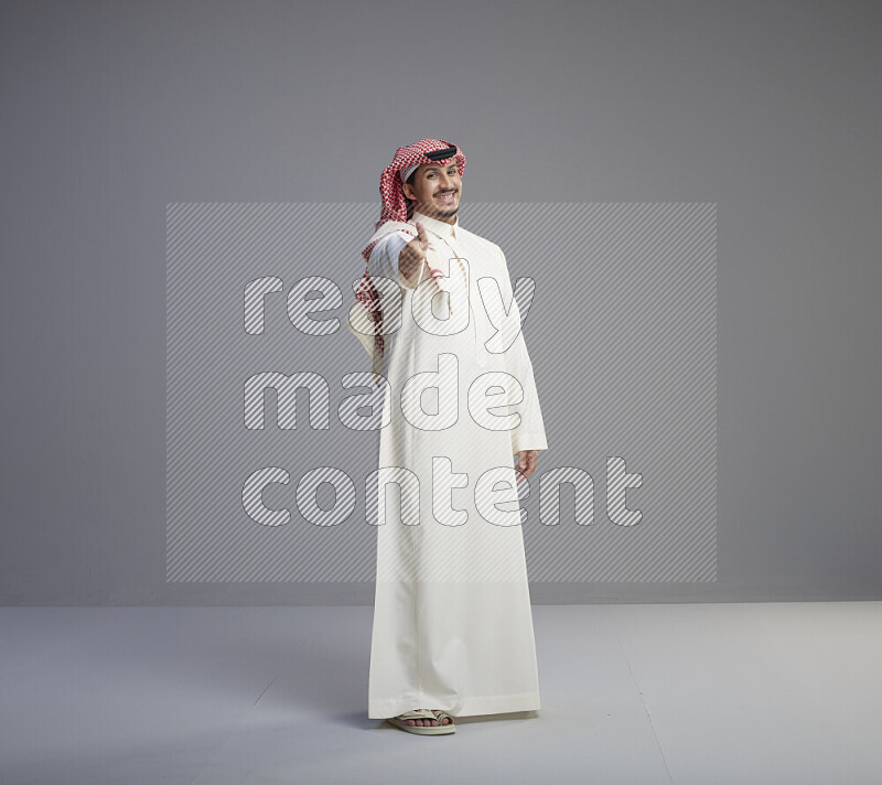 A Saudi man standing wearing thob and red shomag interacting with the camera on gray background