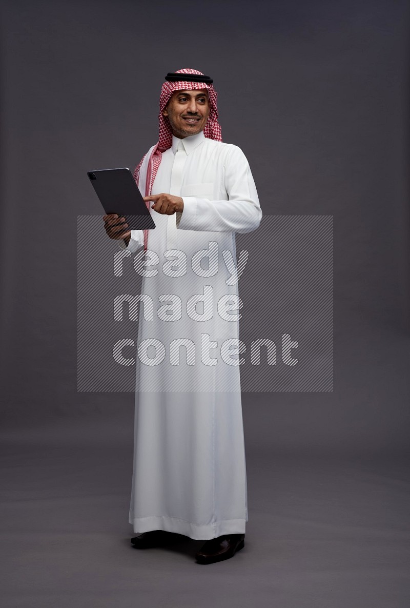 Saudi man wearing thob and shomag standing working on tablet on gray background