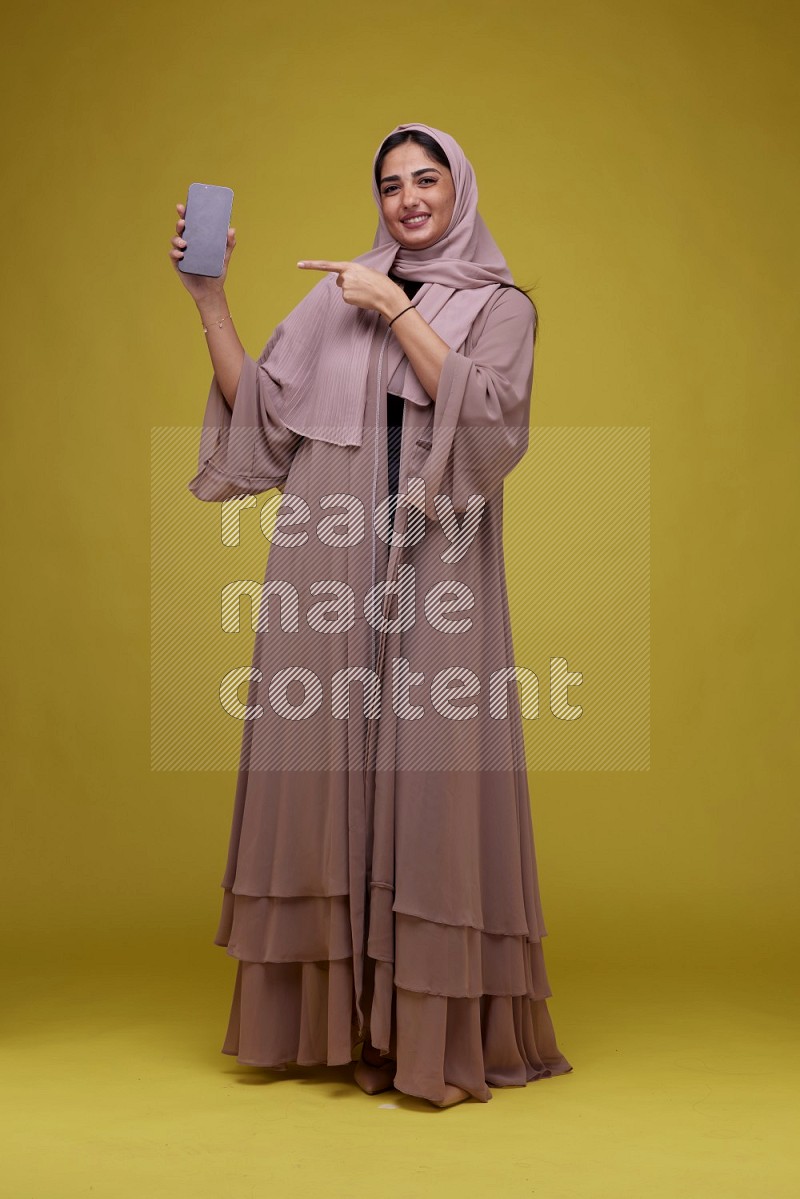 A woman Showing her Phone screen on a Yellow Background wearing Brown Abaya with Hijab