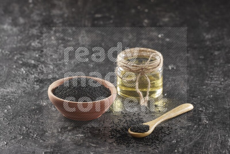 A wooden bowl and spoon full of black seeds and a glass jar of black seeds oil on a textured black flooring