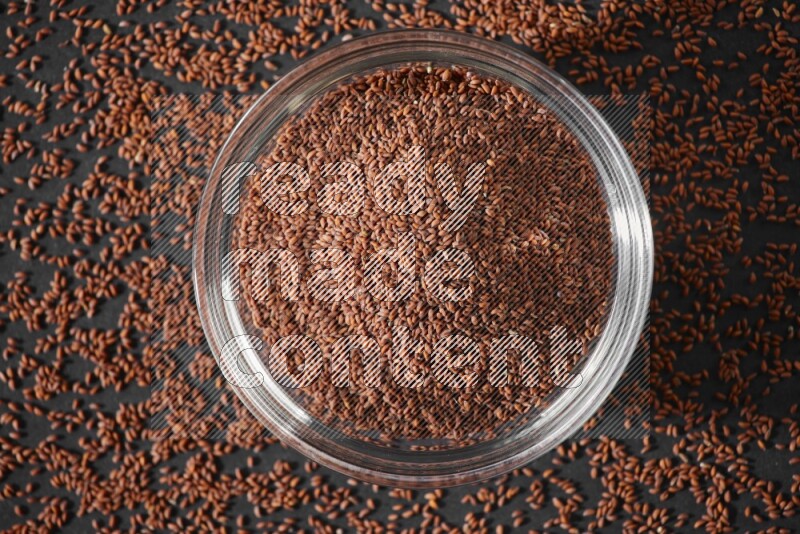 A glass bowl full of garden cress seeds surrounded by seeds on a black flooring