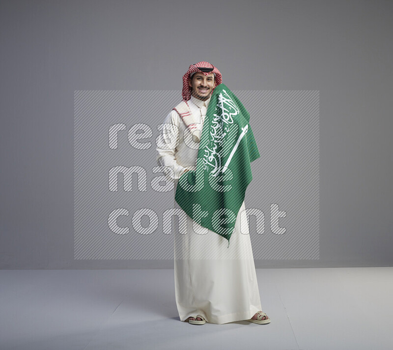 A saudi man standing wearing thob and red shomag holding big saudi flag on gray background