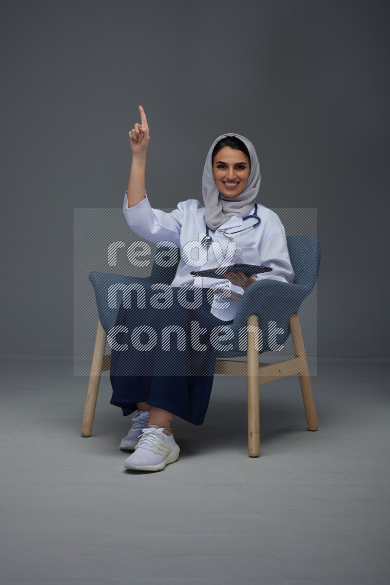 A doctor wearing a light gray head scarf setting on a dark grey chair and using a phone eye level on a grey background