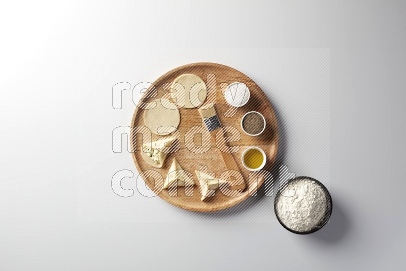 two closed sambosas and one open sambosa filled with cheese while flour, salt, black pepper and oil with oil brush aside in a wooden dish on a white background