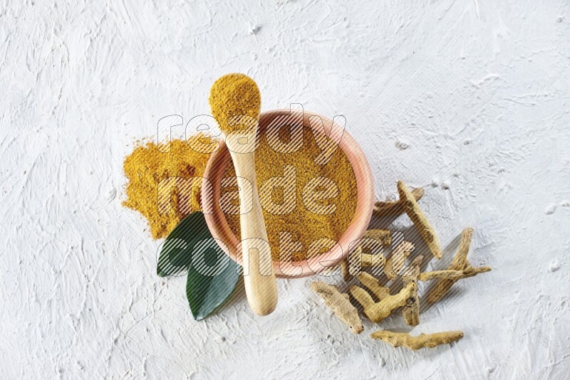 A wooden bowl and wooden spoon full of turmeric powder with dried turmeric fingers beside it on textured white flooring