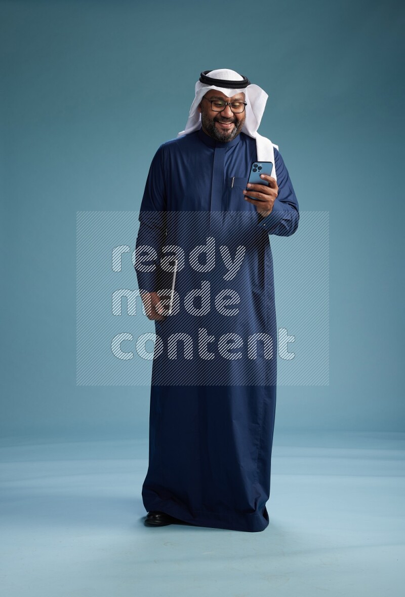 Saudi Man with shimag Standing texting on phone on blue background