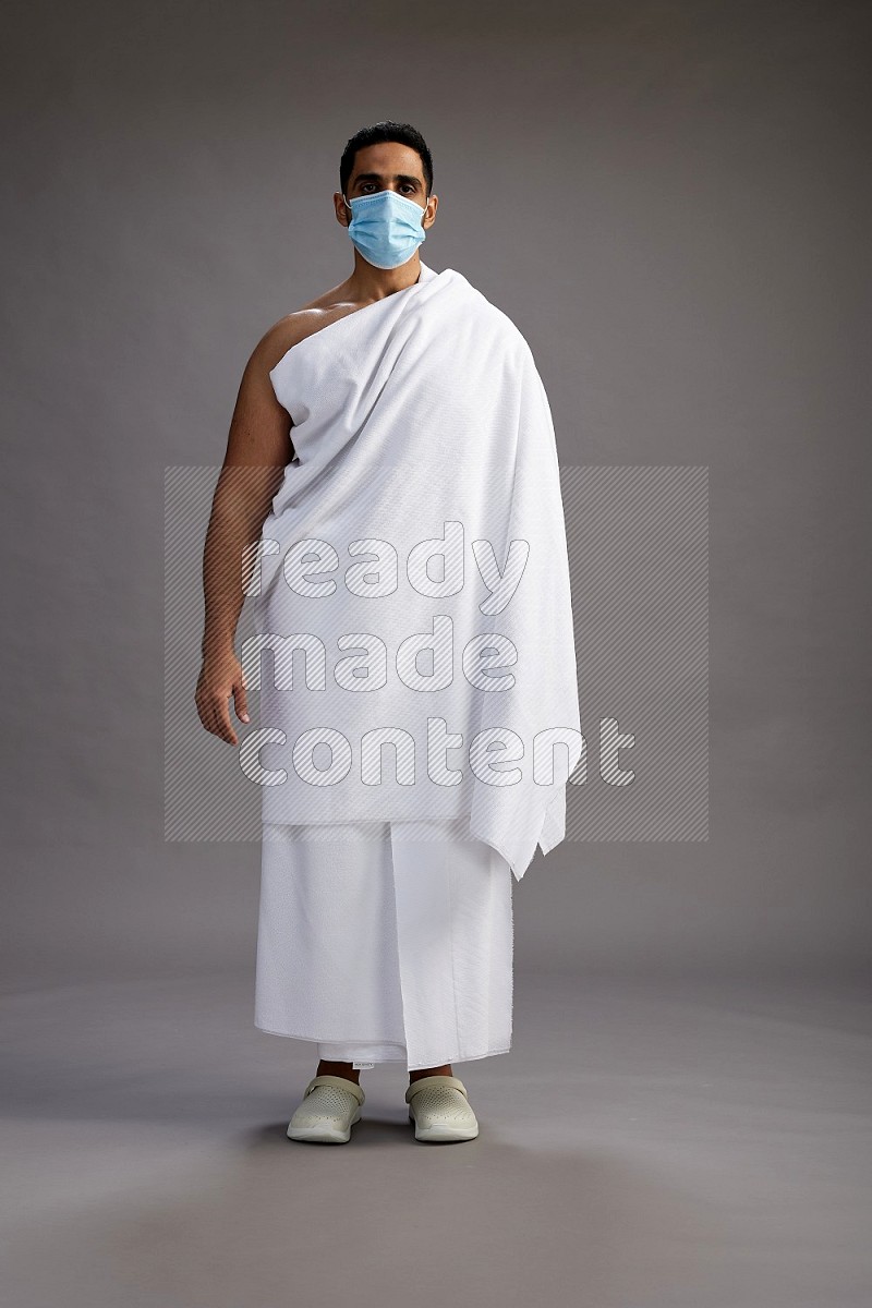 A man wearing Ehram with face mask Standing Interacting with the camera on gray background