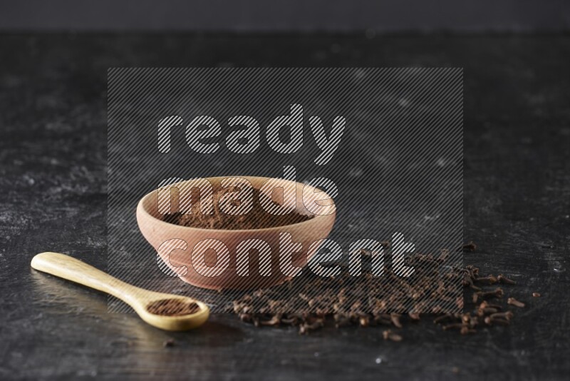 A wooden bowl and wooden spoon full of cloves powder with spreaded cloves on textured black flooring