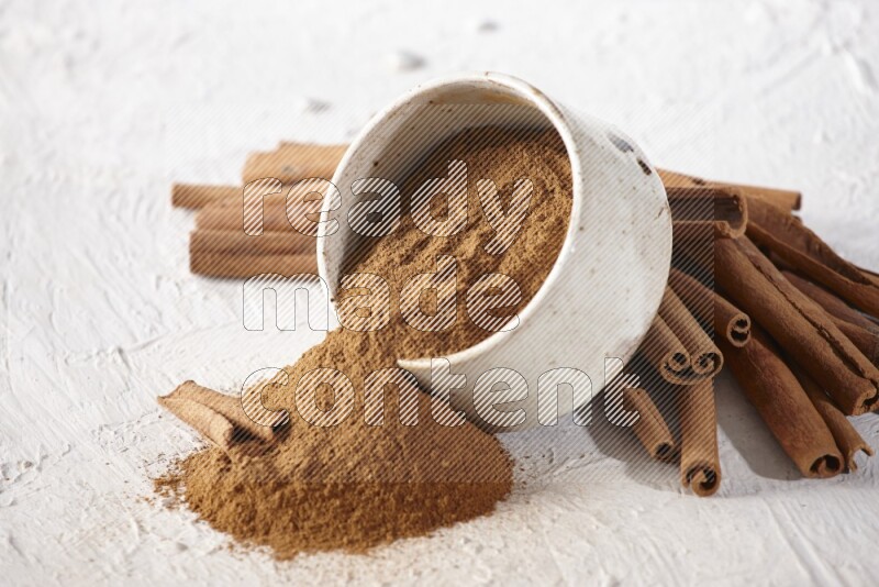 Ceramic beige bowl over filled with cinnamon powder and cinnamon sticks around the bowl on a textured white background in different angles