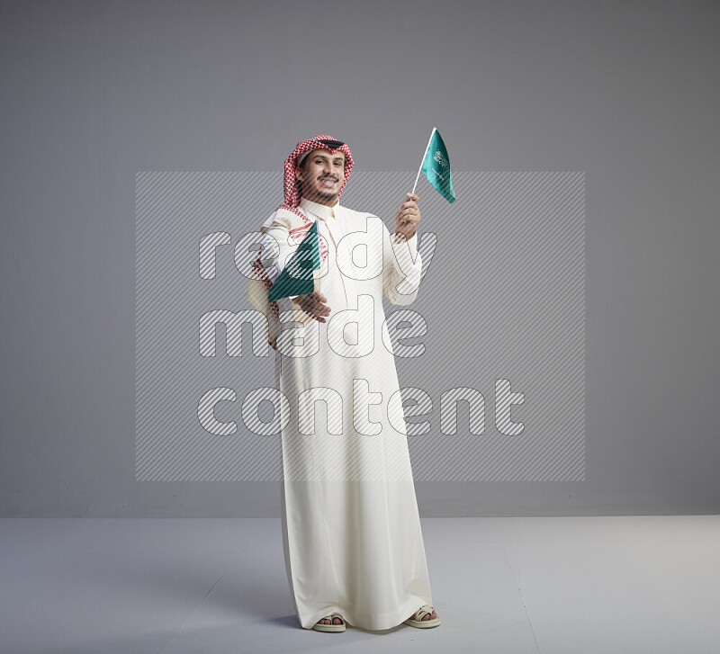 A Saudi man standing wearing thob and red shomag raising small Saudi flag on gray background