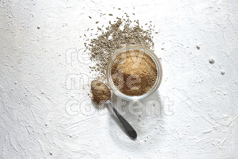 A glass bowl and metal spoon full of cumin powder and cumin seeds underneath it on textured white flooring