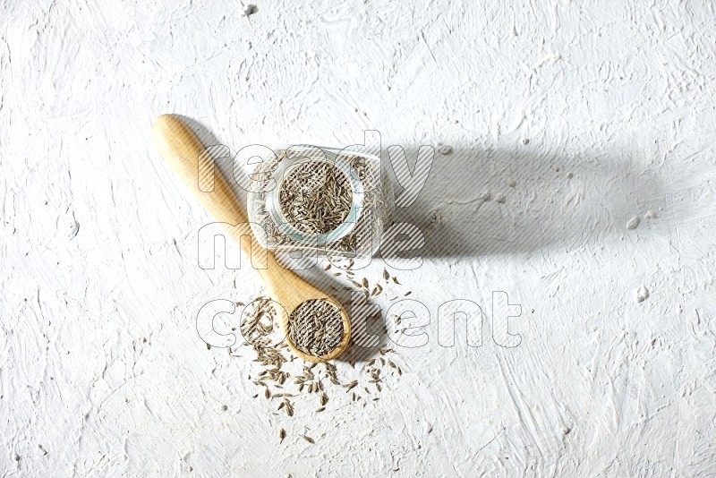 A glass spice jar and wooden spoon full of cumin seeds on textured white flooring