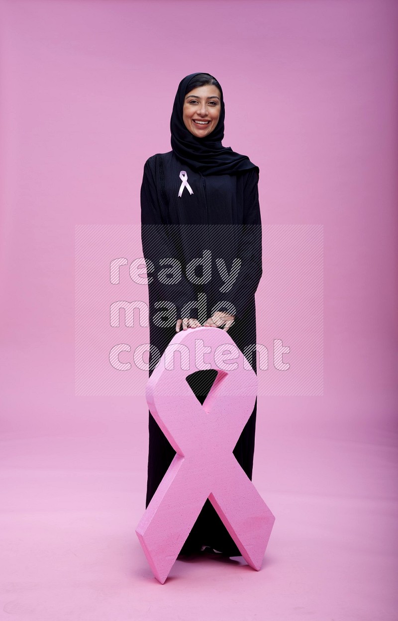 Saudi woman wearing pink ribbon on abaya standing holding awareness ribbon on plain pink background