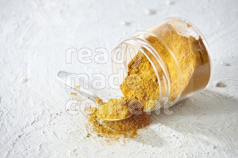A flipped glass spice jar and a metal spoon full of turmeric powder and powder spilled out of it on textured white flooring