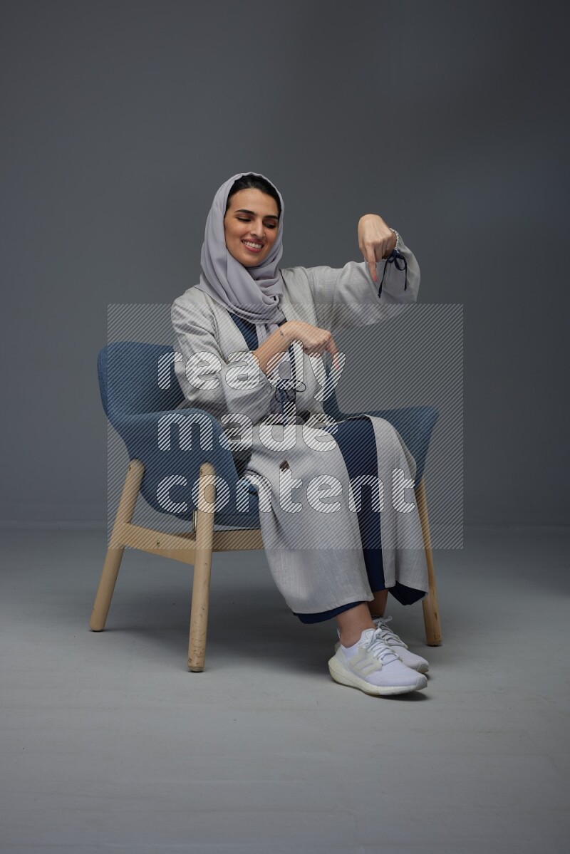 A Saudi woman wearing a light gray Abaya and head scarf sitting on a dark grey chair while making multi hand gestures eye level on a grey background
