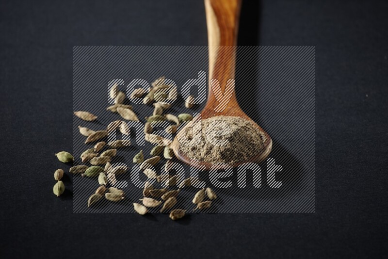 A wooden ladle full of cardamom powder and cardamom seeds beside it on black flooring