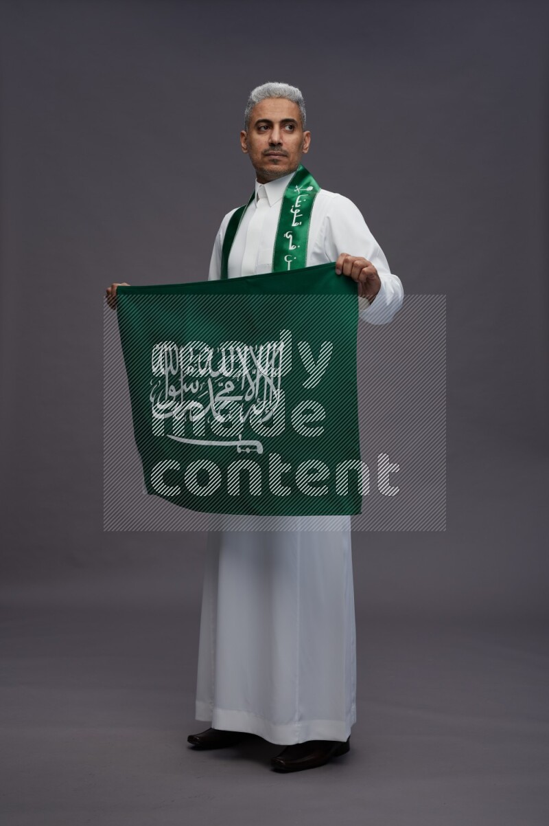 Saudi man wearing thob standing holding Saudi flag on gray background