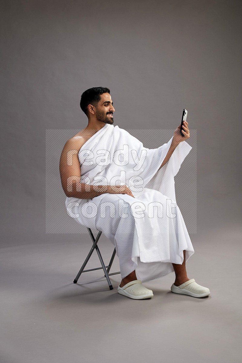 A man wearing Ehram sitting on chair taking selfie on gray background