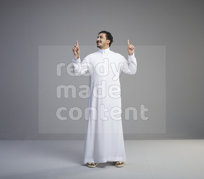 A Saudi man standing wearing thob with face painting interacting with the camera on gray background