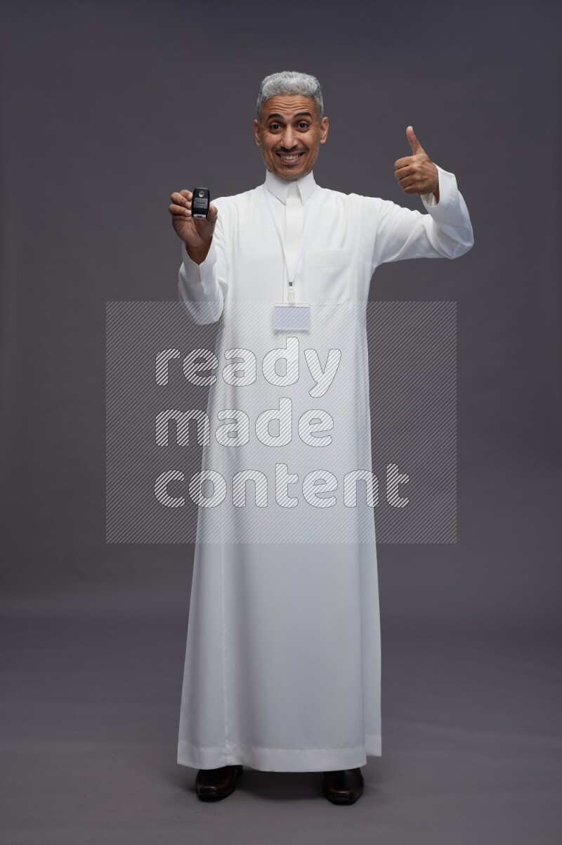 Saudi man wearing thob with neck strap employee badge standing holding key car on gray background