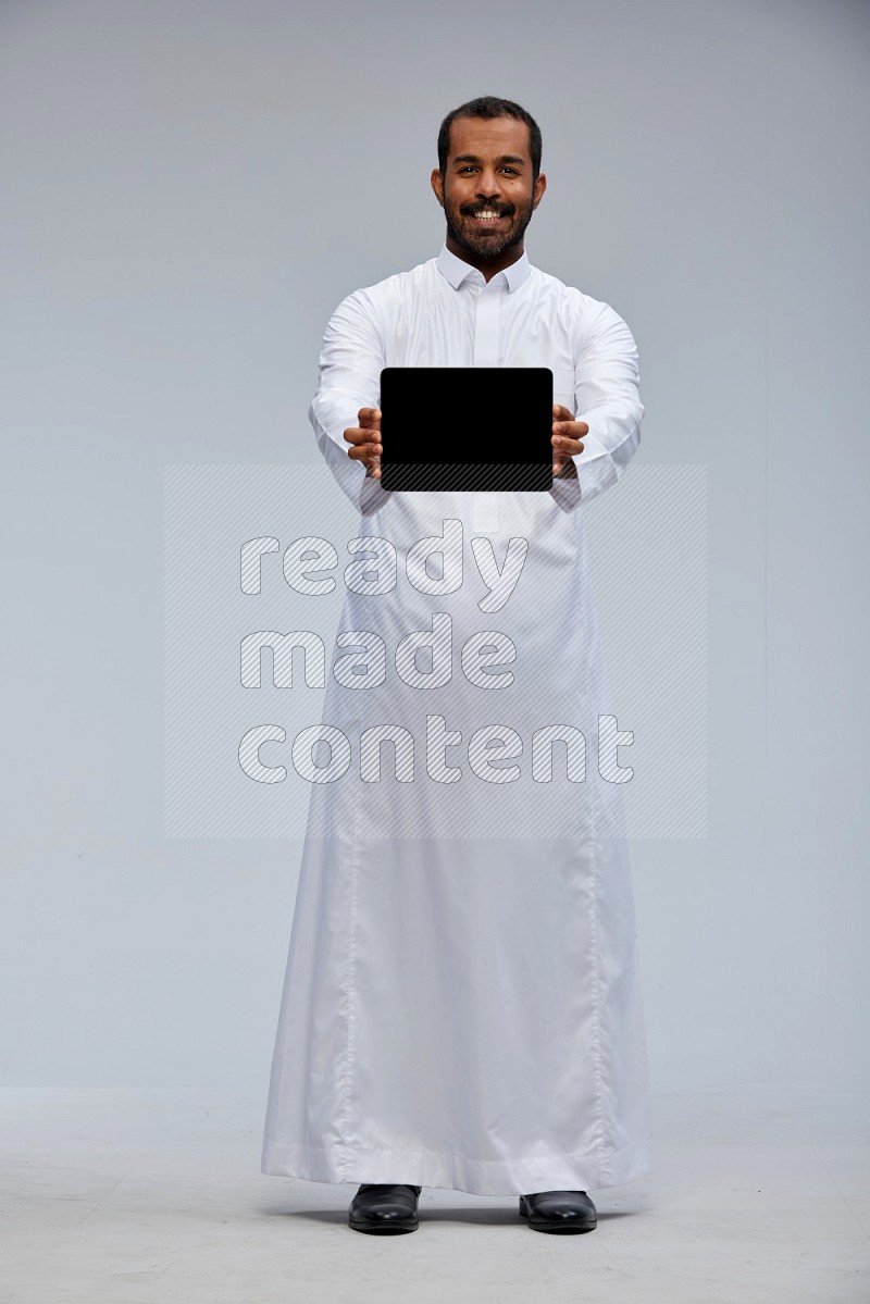 Saudi man wearing Thob standing showing tablet to camera on Gray background