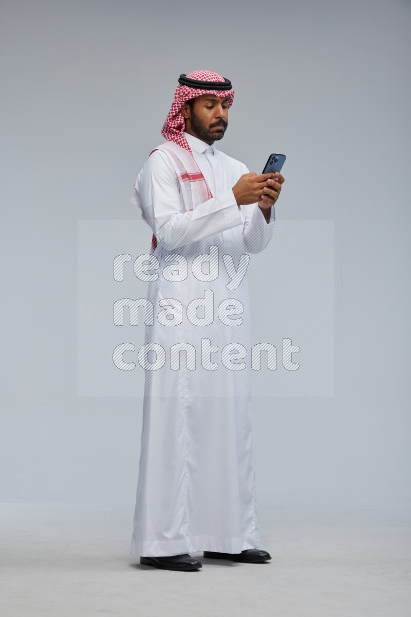Saudi man Wearing Thob and shomag standing texting on phone on Gray background